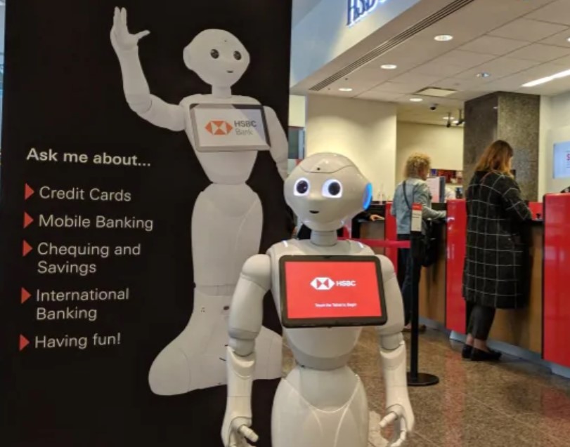 A robot standing next to a sign advertising a bank. The robot has a tablet on its chest.