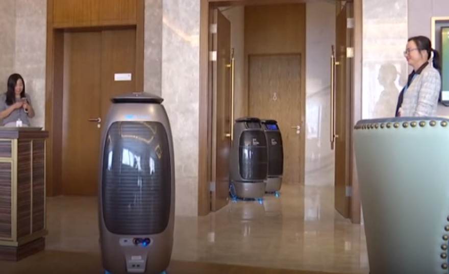 A modern lobby with people standing, two robotic vacuum cleaners, a reception desk, and an elevator.