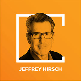 A man in a suit, smiling at the camera. He has grey hair and glasses. The background is orange with a white square frame around him.