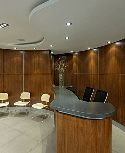 An interior view of an office lobby, featuring a modern reception desk with chairs, a large window, and a wood paneled wall.