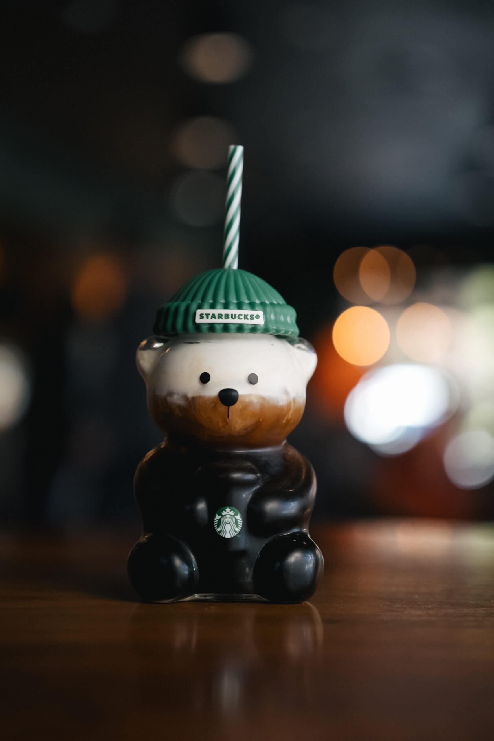 A small teddy bear figurine wearing a green hat, sitting in front of a cupcake on a table.