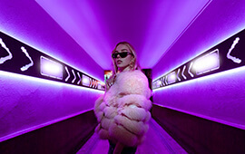 A woman wearing sunglasses and a fur coat, standing in front of a purple light tunnel.