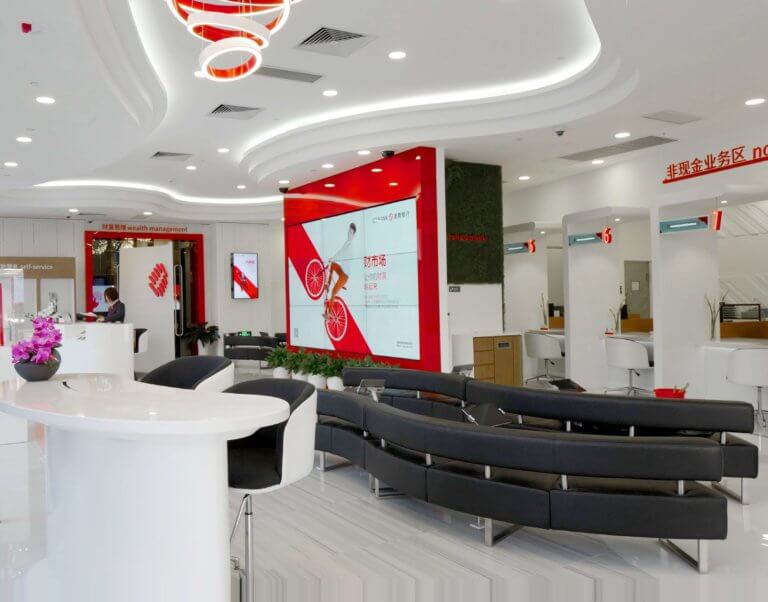 A modern, well-lit lobby with a large flat screen tv displaying red and white colors, a reception desk, seating area with black chairs, and a sign…