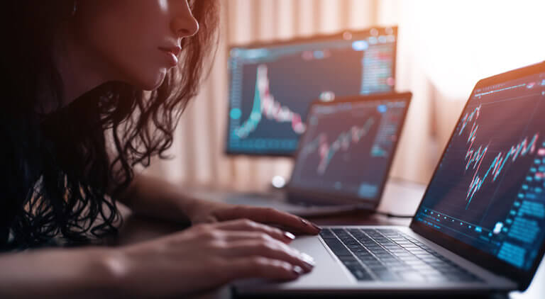A person sitting at a desk working on a laptop, surrounded by multiple computer monitors displaying various charts and graphs.