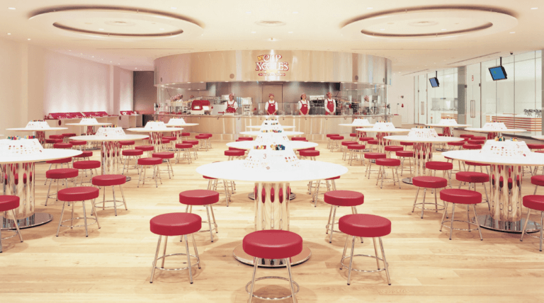 Empty restaurant interior with round tables, red chairs, and a large open kitchen area.