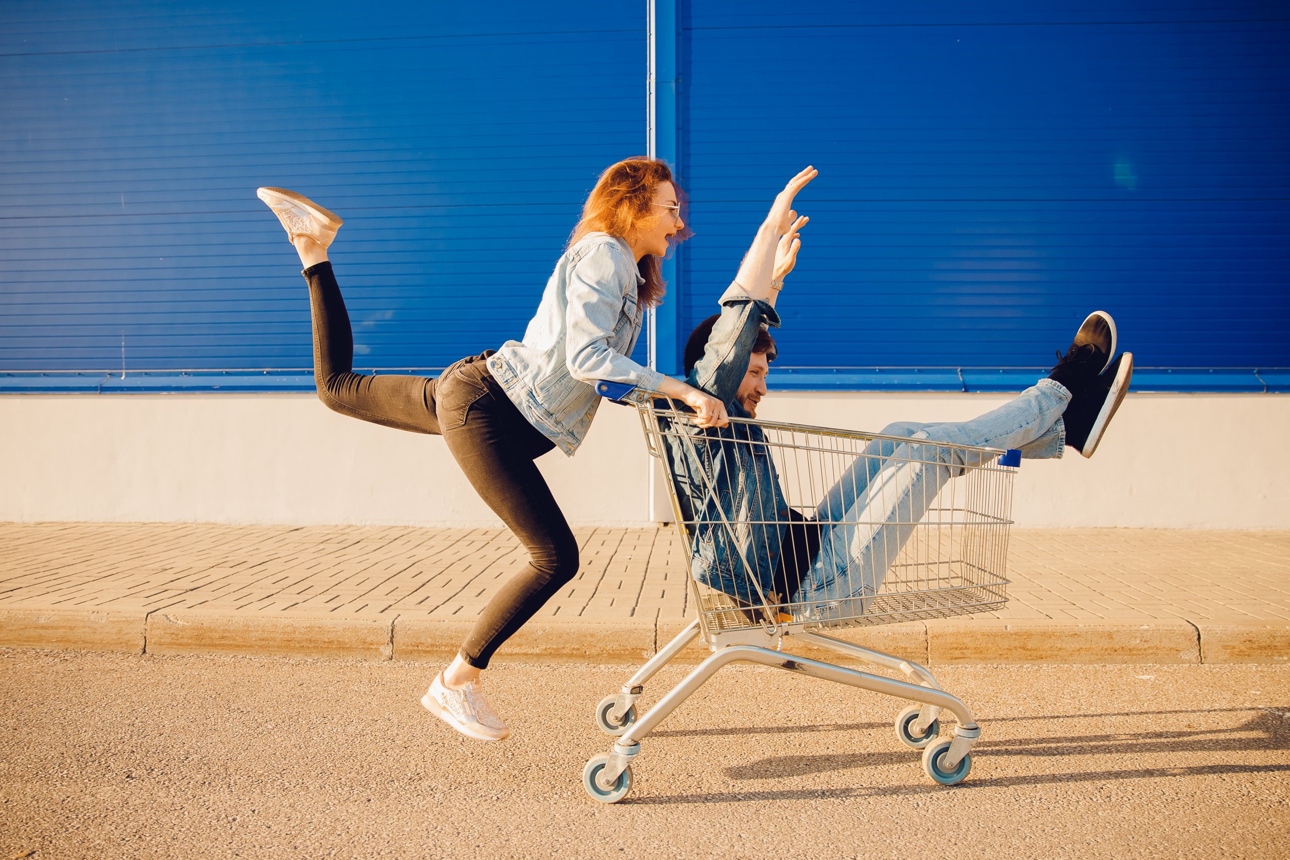 Two people playfully posing on a shopping cart, one person in the air and another on the ground, both holding onto the cart.