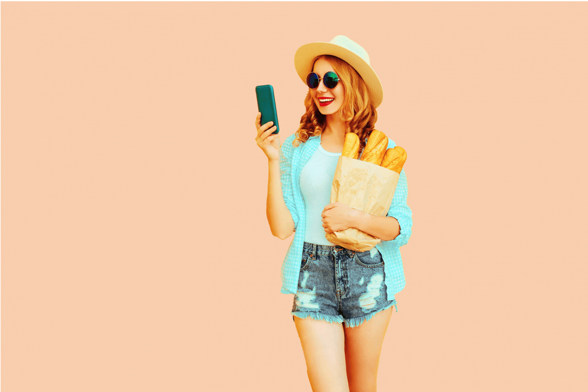 Woman holding phone, wearing sunglasses, straw hat, denim shorts, standing against pink background.
