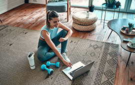 A woman sitting on the floor, using her laptop, surrounded by exercise equipment.
