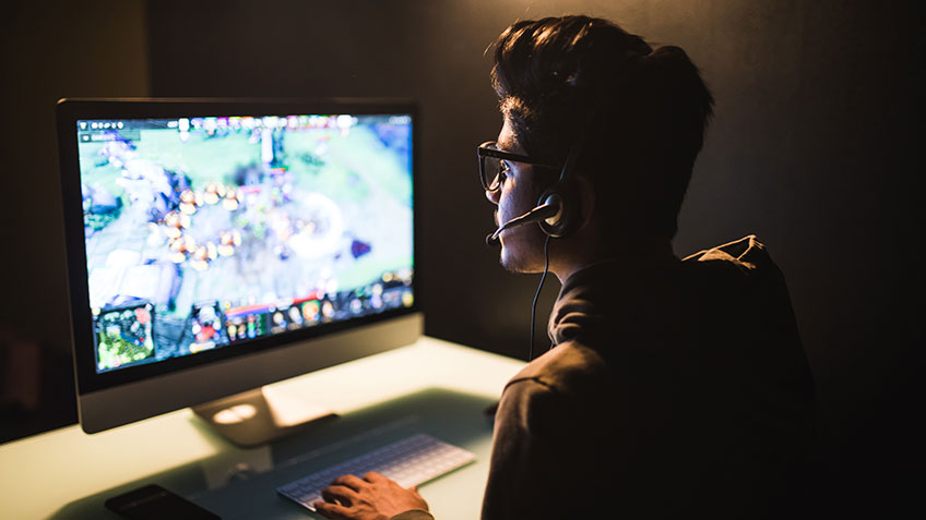 A person sitting at a desk playing video games, wearing headphones and glasses, in front of a large monitor displaying the game.
