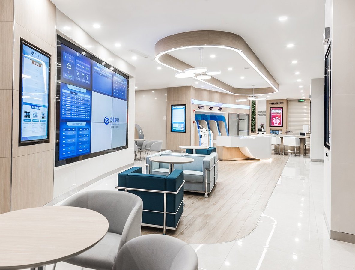 A modern, well-lit lobby with a large screen displaying information, comfortable seating areas, and a reception desk.
