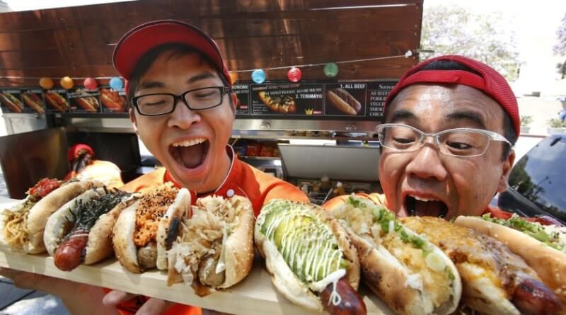 Two men posing with hot dogs, one holding a tray of assorted food items.