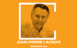A square graphic with an orange background and a white border. Inside the square, there's a photograph of a man wearing glasses and a dark suit.…
