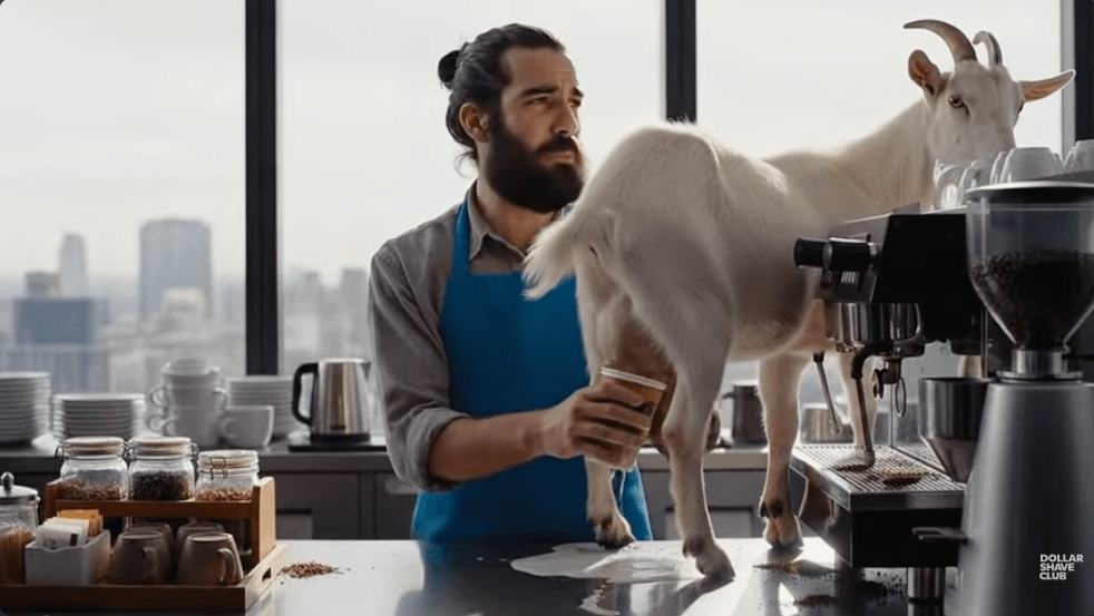 A man standing behind a counter with a goat on the counter, both in front of a coffee machine. The man is milking the goat into a coffee cup.