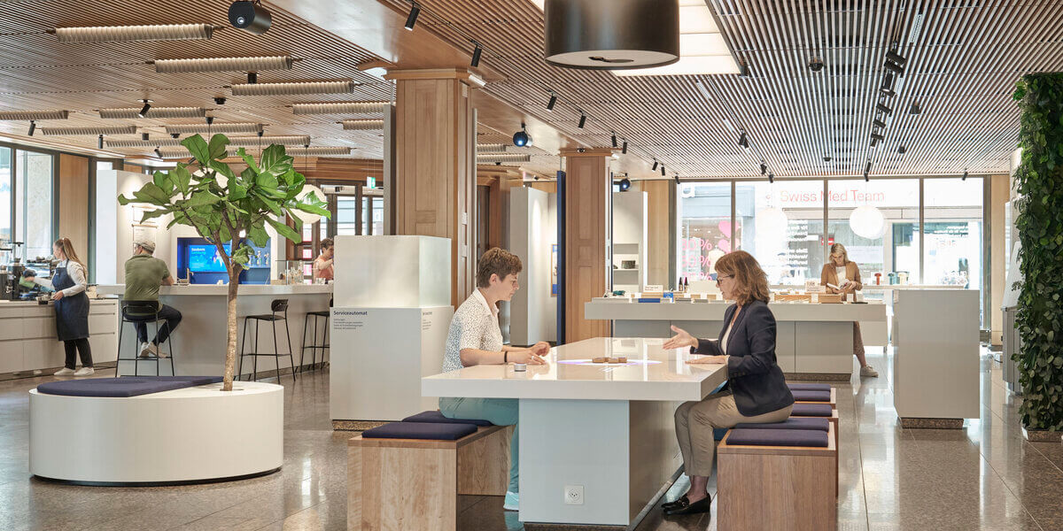 A modern interior space, possibly a lobby or waiting area, with two people seated at a table, surrounded by contemporary furniture and decor.