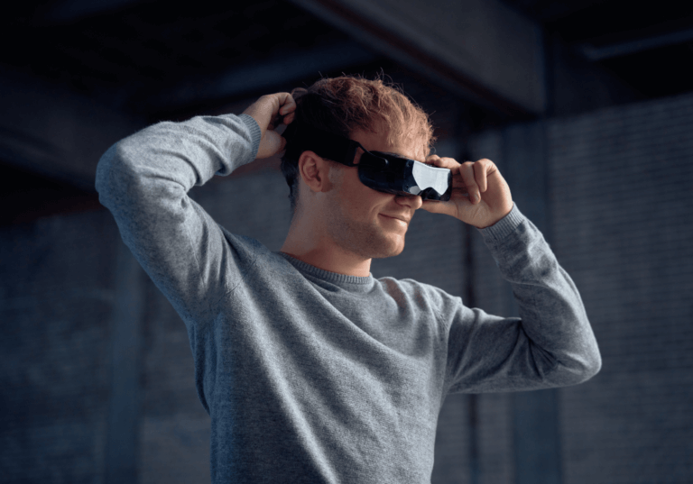 Man in grey sweater holding goggles over eyes, standing against brick wall.