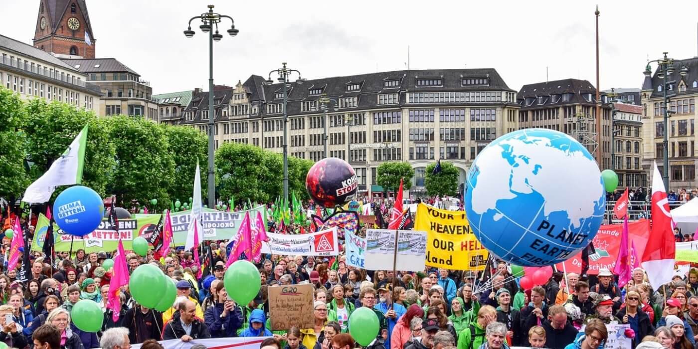 A large crowd of people gathered on a street, holding signs and balloons, protesting in front of a building.