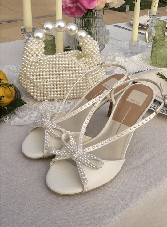 A pair of white sandals with pearls and a bow on a table, next to a handbag.