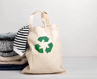 A white tote bag with a green recycling symbol on it, placed next to a pile of laundry on a bed.
