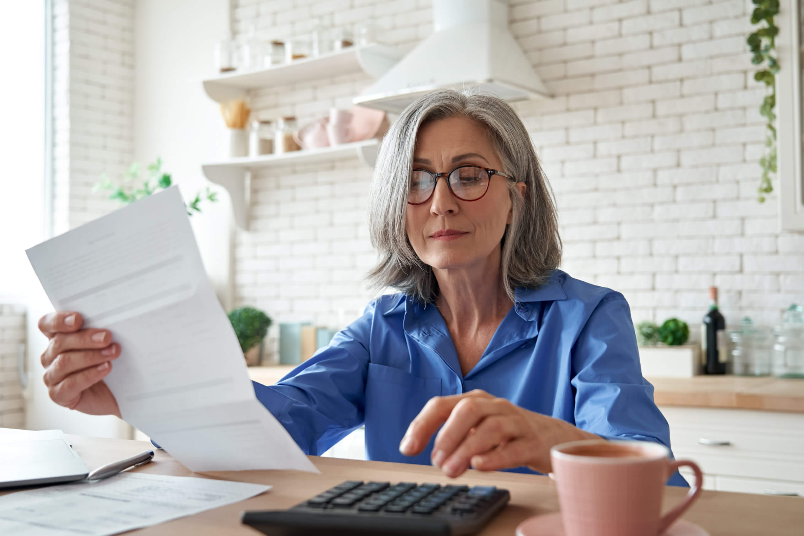 Woman sitting at kitchen table, reading paperwork, wearing glasses, blue shirt, and holding pen.
