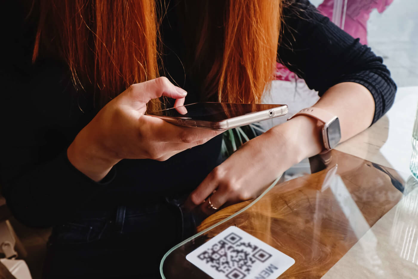 A woman sitting at a table, looking at her phone. She has red hair and is wearing a black shirt.