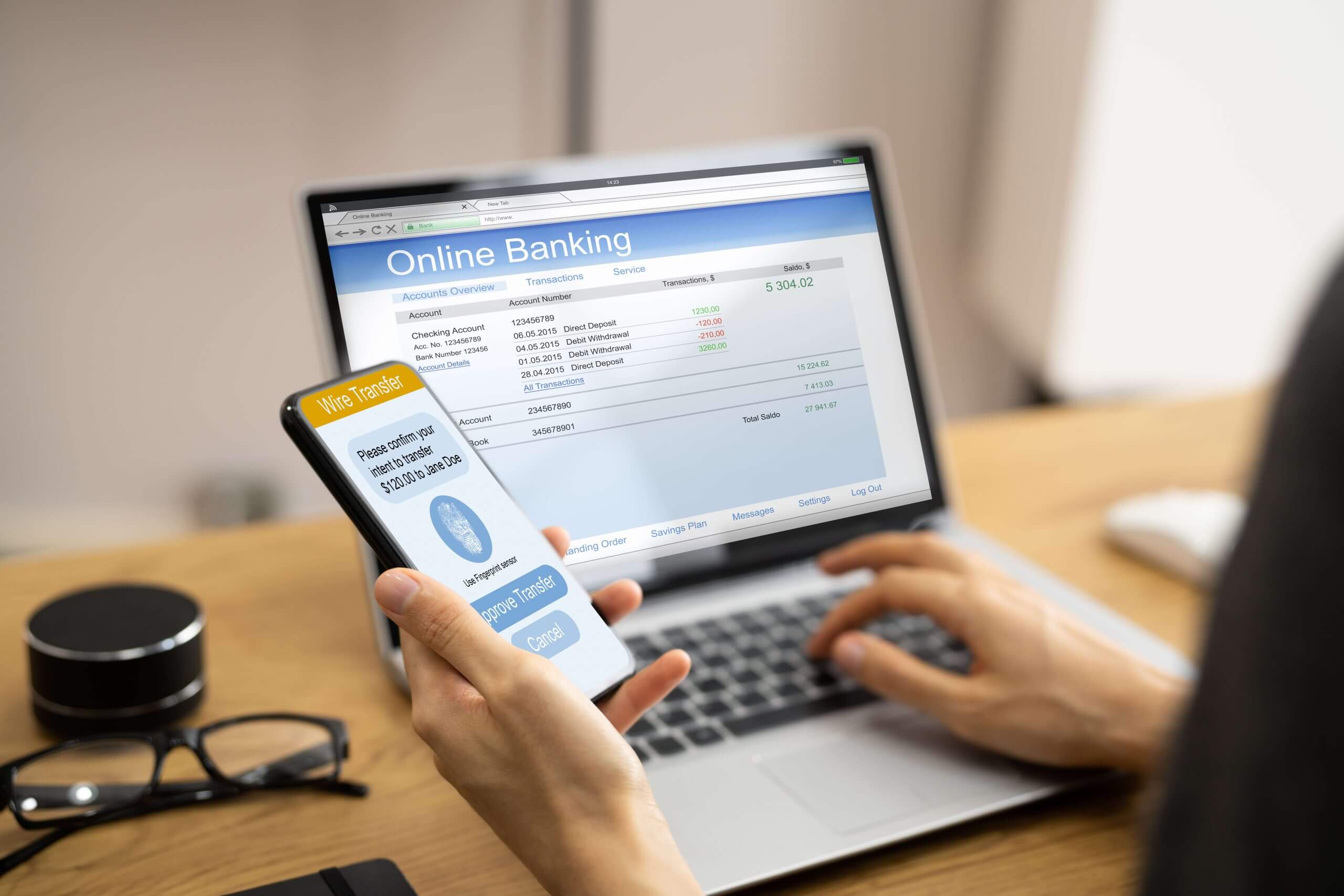 A person holding a smartphone displaying a banking app, showing an account balance and transaction details on a laptop screen in the background.