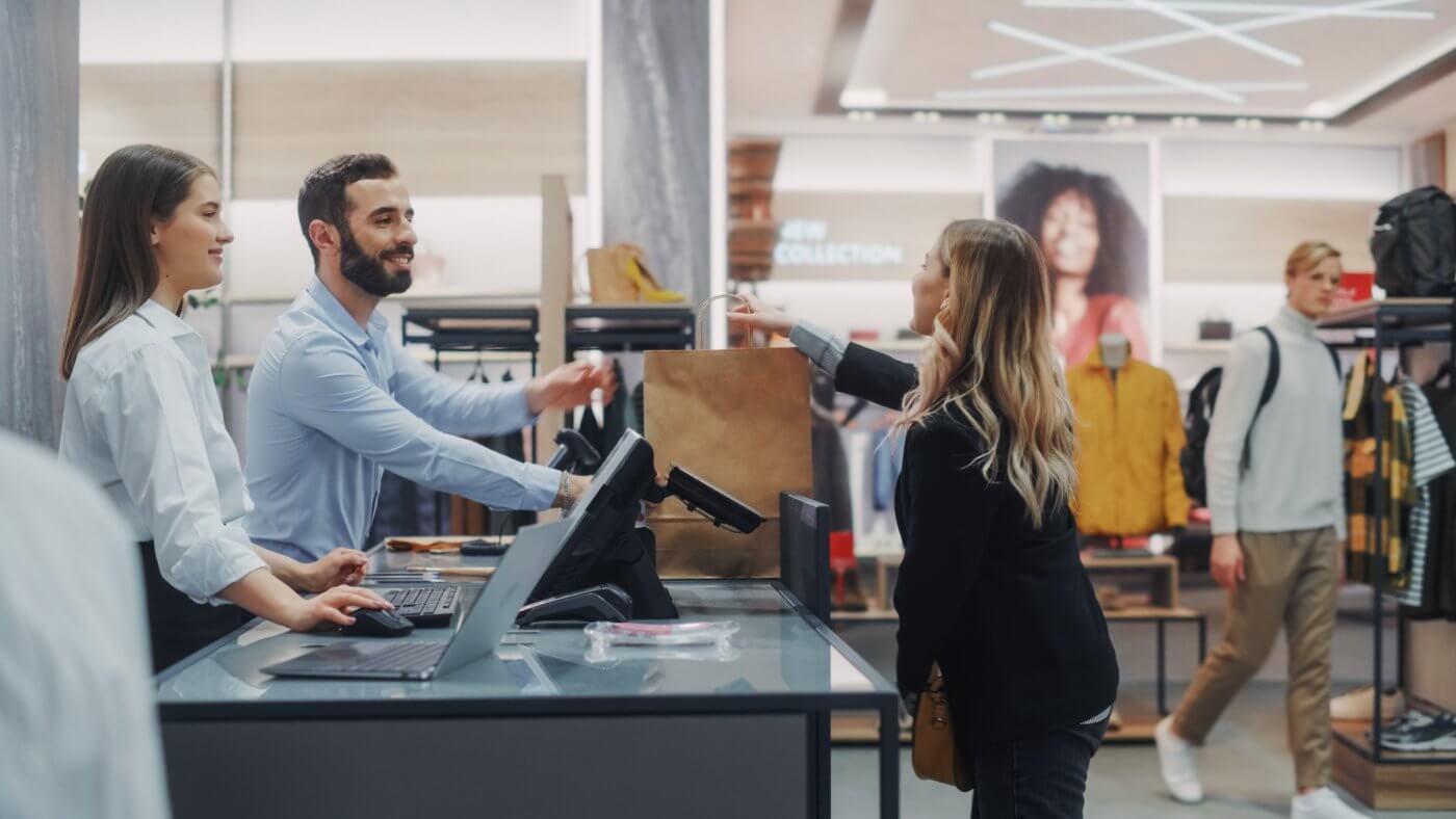 An image of a store scene, featuring two people standing behind a counter, one of whom appears to be a salesperson, and another person in the…