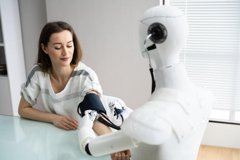 Woman sitting at desk with robotic arm, both looking at it.