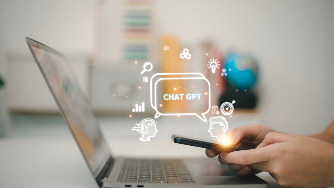 A laptop screen with a chatbot interface, featuring a speech bubble icon and a person's hand holding a smartphone.