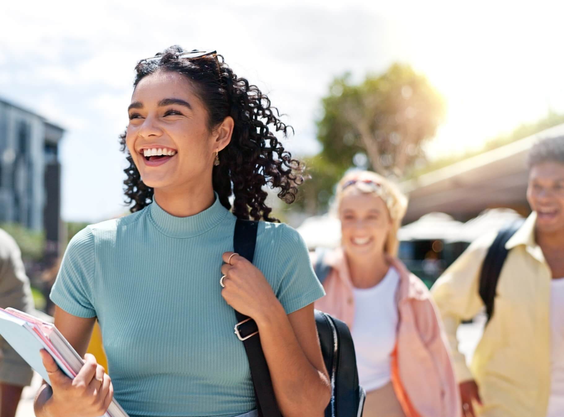 A young woman smiling, holding a book, walking down the street with other people.