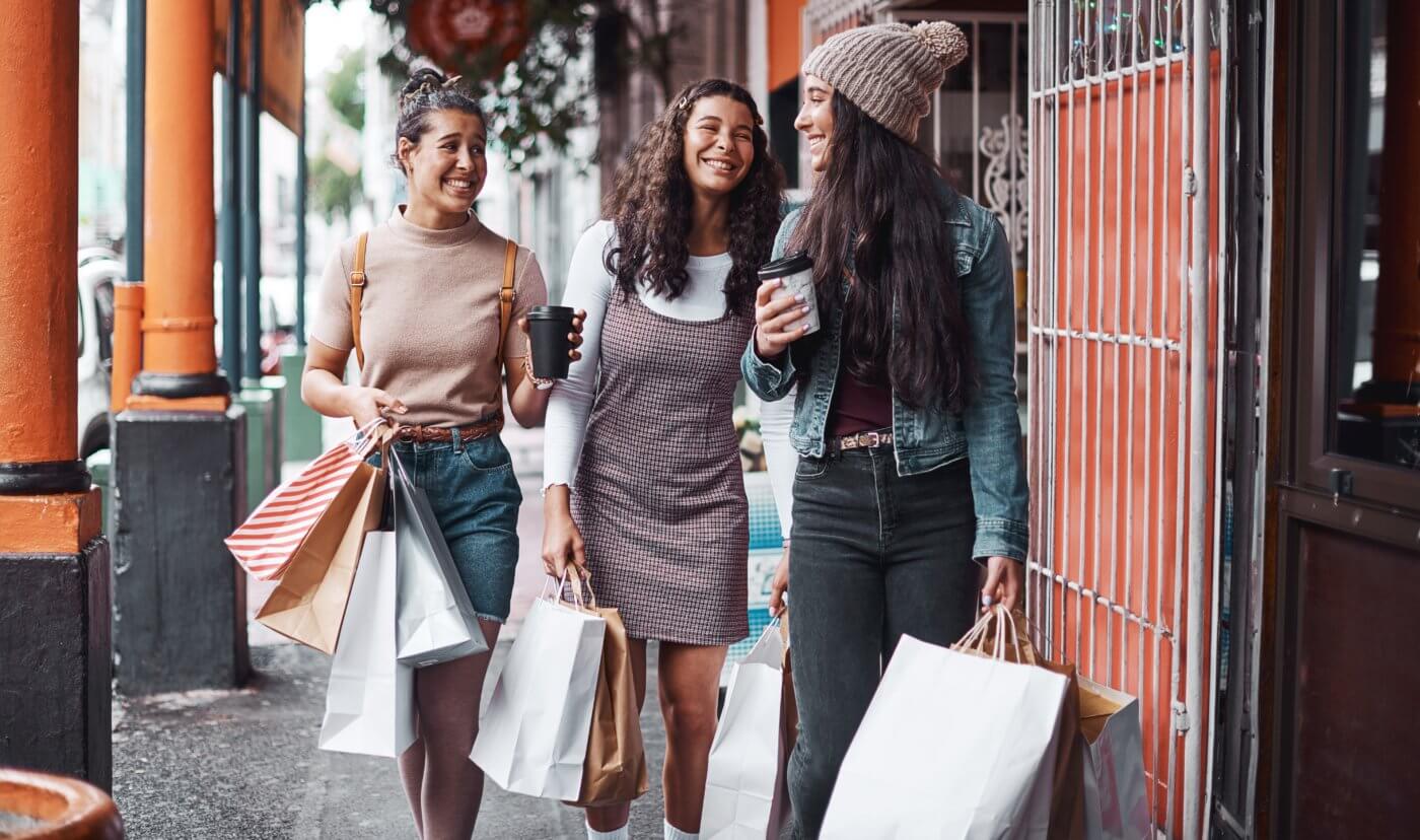 Three people walking on sidewalk, smiling, carrying shopping bags.
