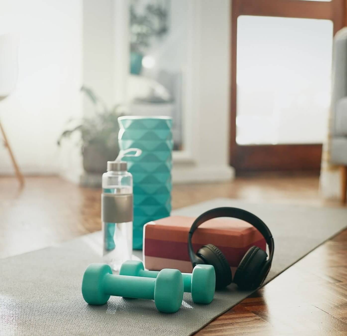 A living room setting with exercise equipment, including dumbbells, a yoga mat, and a water bottle, on a wooden floor next to a window.