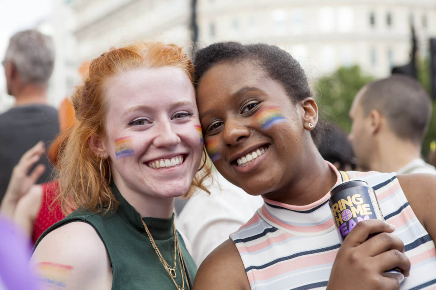 Two women standing together, smiling at the camera. One woman has a rainbow flag painted on her face. They are holding a sign that says