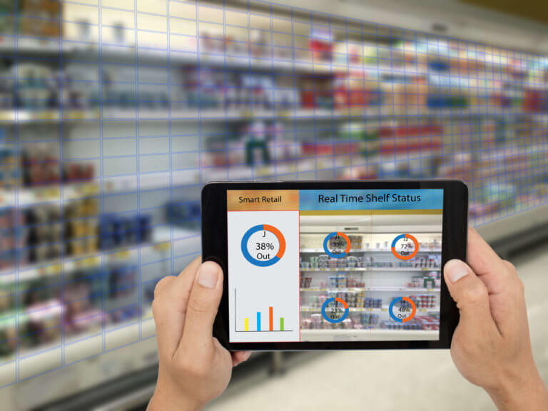 A person holding a tablet displaying a dashboard with various statistics, such as sales data, on a screen in front of a grocery store shelf…