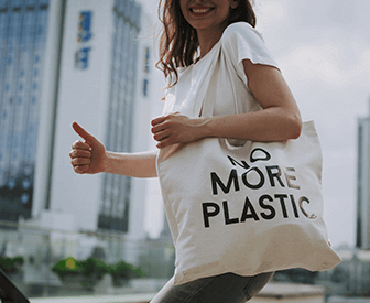 A woman holding a white tote bag with the words "more plastic" on it, giving a thumbs up.